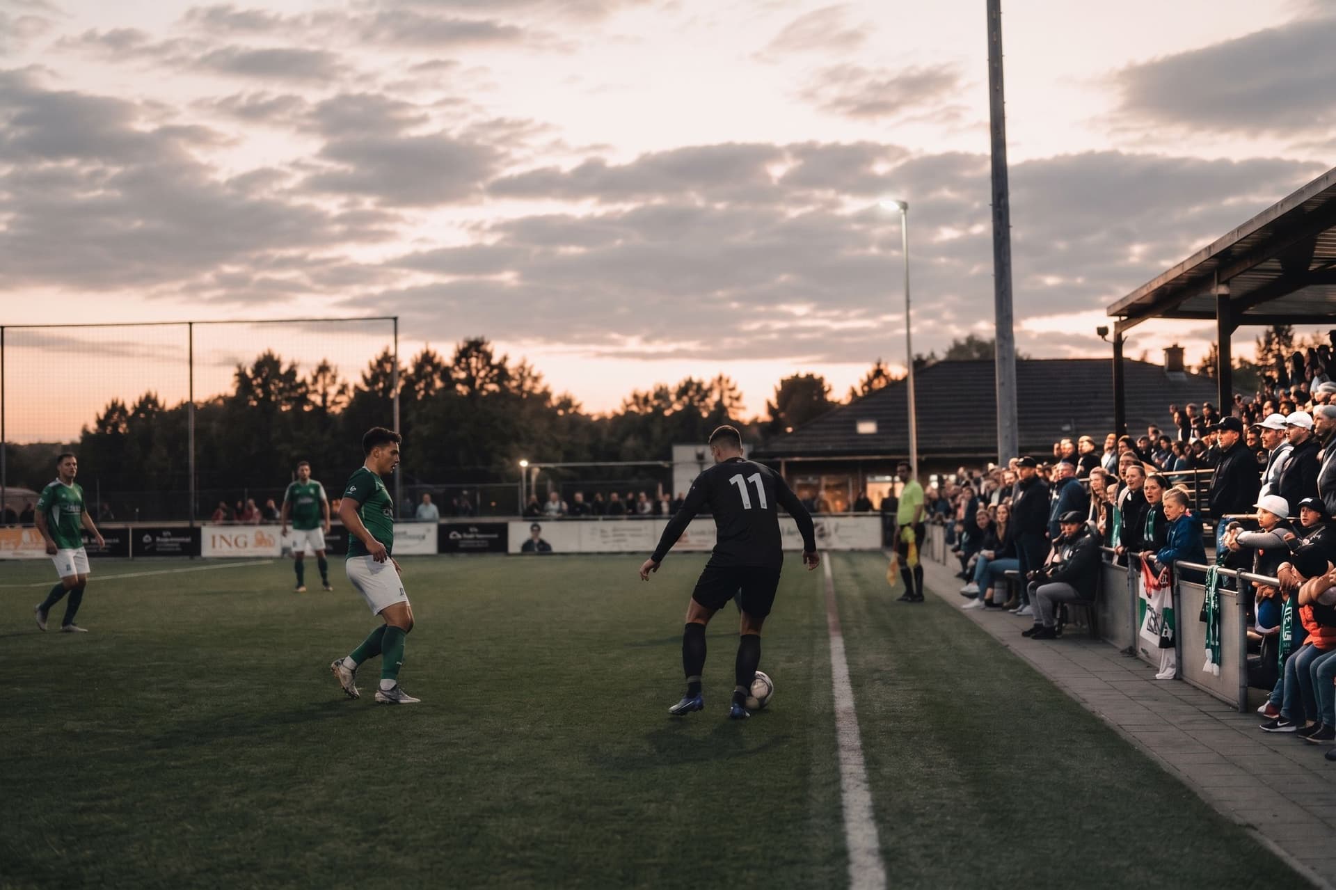 Avondwedstrijd bij een amateurclub, met publiek en sponsorborden langs de lijn
